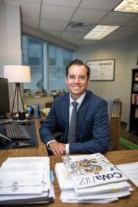 Smiling man in a blue suit sits at a wooden desk in a bright office, with papers and magazines in the foreground.
