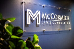 Acrylic sign on a blue wall reading 'MCCORMICK LAW & CONSULTING' with a white geometric logo, foreground plant leaves partially obscuring the view.