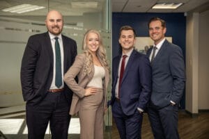 Four professionals in business attire posing together in a modern office lobby with glass partition.