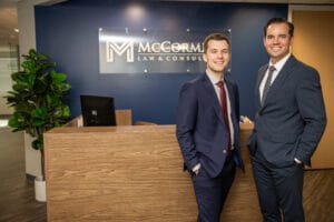 Two men in business suits standing at a wooden reception desk in a law office, blue wall behind them with a glass sign.